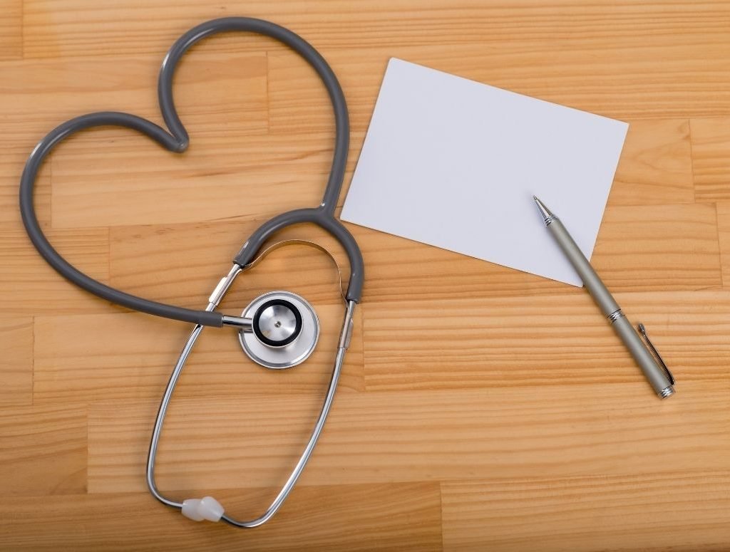Image of a stethoscope, a black prescription pad, and a pen on a wooden table