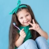 Image of a smiling young girl holding a toothbrush and making the peace sign