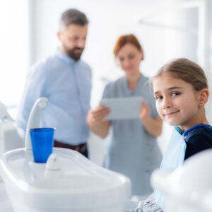 Image of a child smiling at you while two adults, one of which is a dental practitioner, are looking at presumably her diagnosis in a blurred foreground