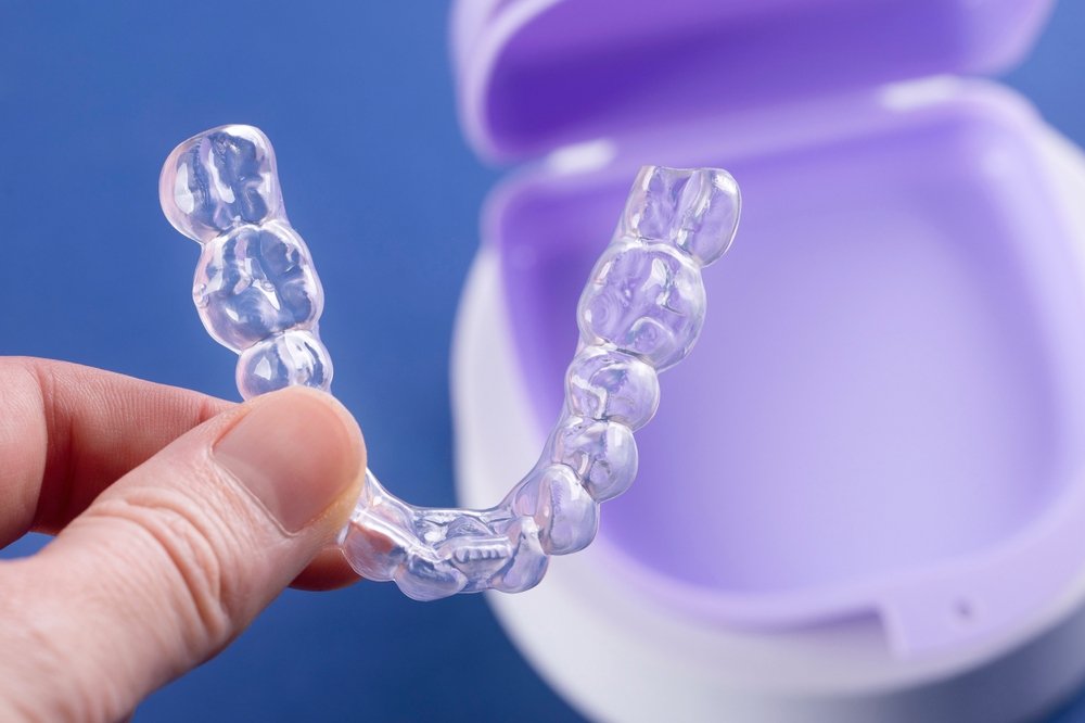 A hand holds a Clear Aligners tray in front of an open purple and white storage case on a blue background.