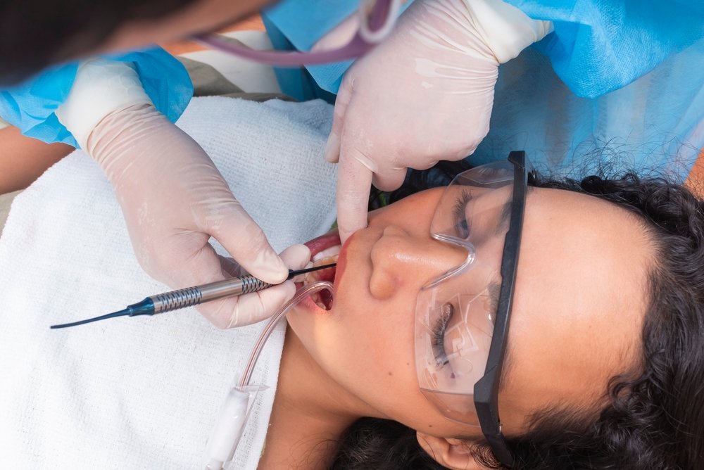 A dentist wearing gloves and a protective gown works on a patient's teeth. The patient lies back wearing safety glasses and a bib, while the dentist uses dental instruments inside the patient's mouth.