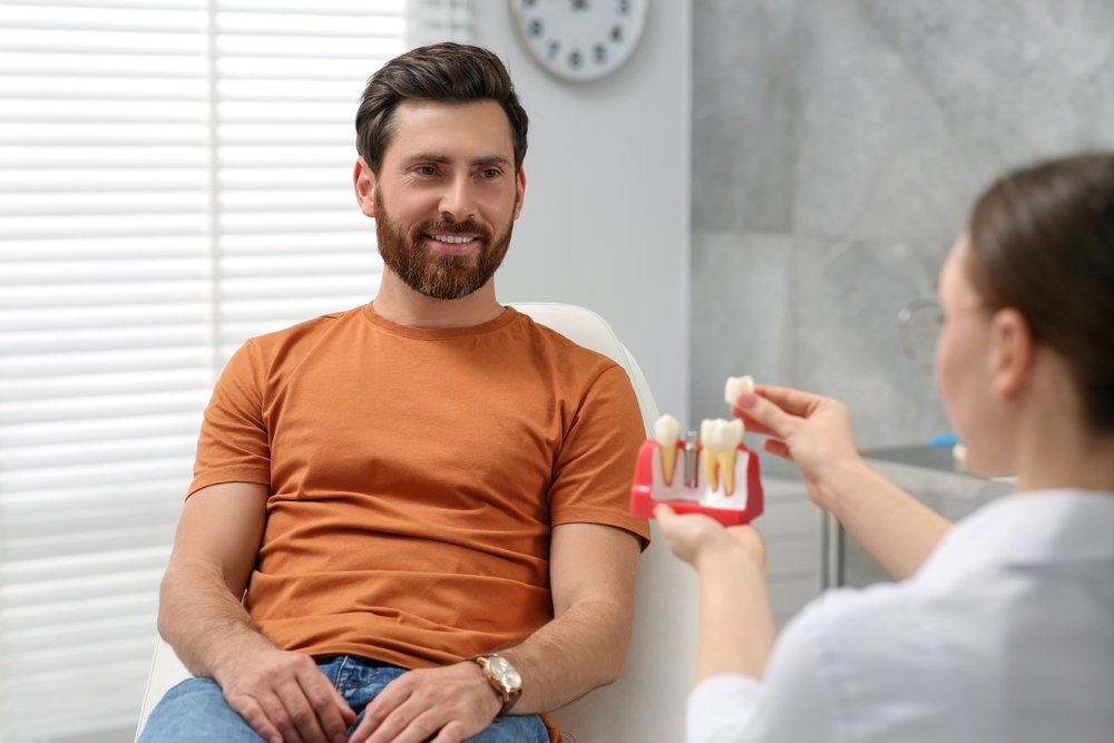 A man sits in a dental surgery, smiling at a dental professional who is showing him a dental implant model.