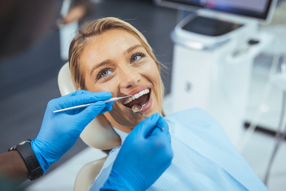 A woman sits in a dental chair, smiling with her mouth open as a dentist wearing blue gloves examines her teeth with dental instruments.