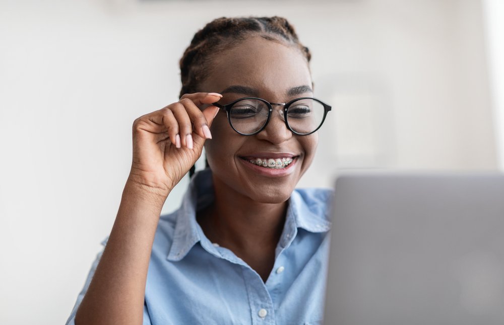 A smiling woman with braces wearing glasses looks at a laptop.
