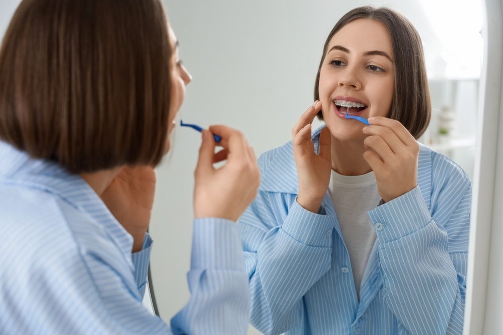 An adult with braces using an interdental brush to clean their teeth in a mirror.